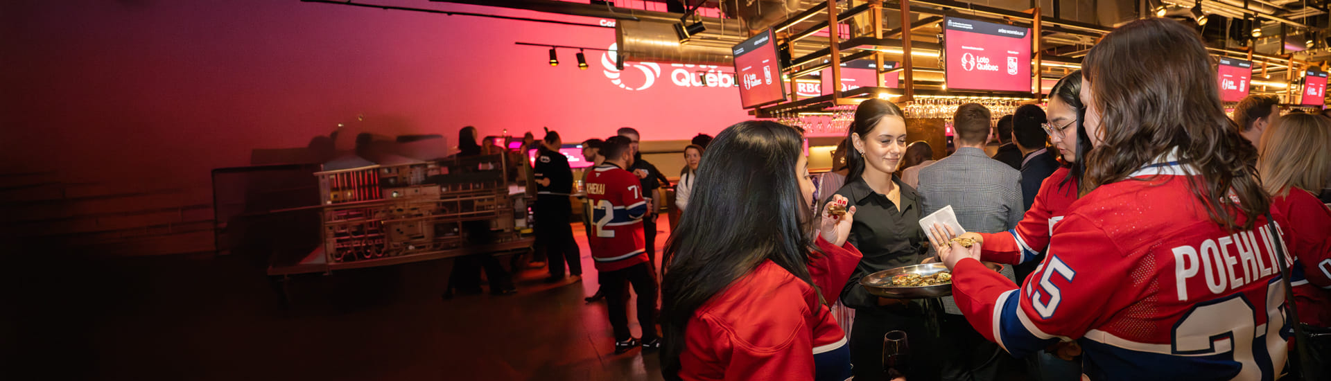 Participants at a networking event in a reception area; several people wearing hockey jerseys are interacting with guests, while one person is serving appetizers. Screens displaying partner logos are visible in the background.