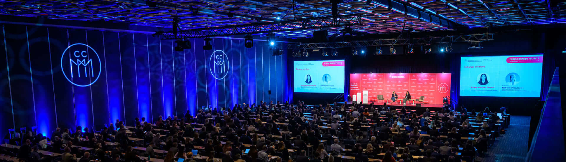Overview of a Chamber of Commerce of Metropolitan Montreal conference; numerous attendees are gathered in a large room facing a stage where a discussion is taking place, with presentation screens and blue and red lighting.