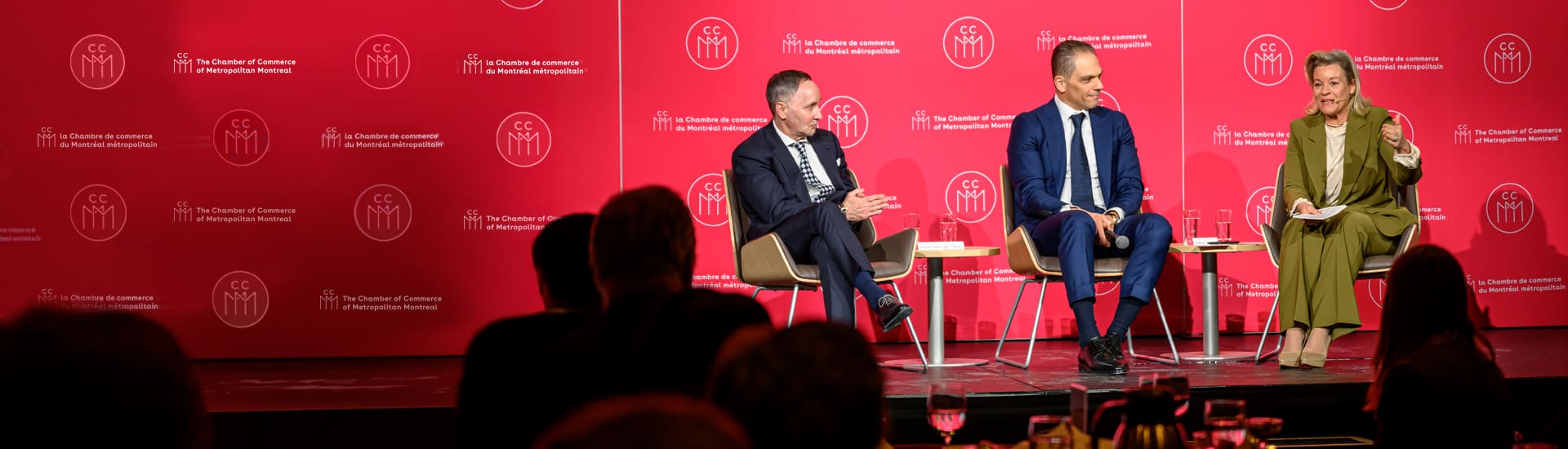 Panel discussion on stage at a Chamber of Commerce of Metropolitan Montreal event; several panelists are participating in a conversation before an audience, against a red backdrop featuring CCMM branding.