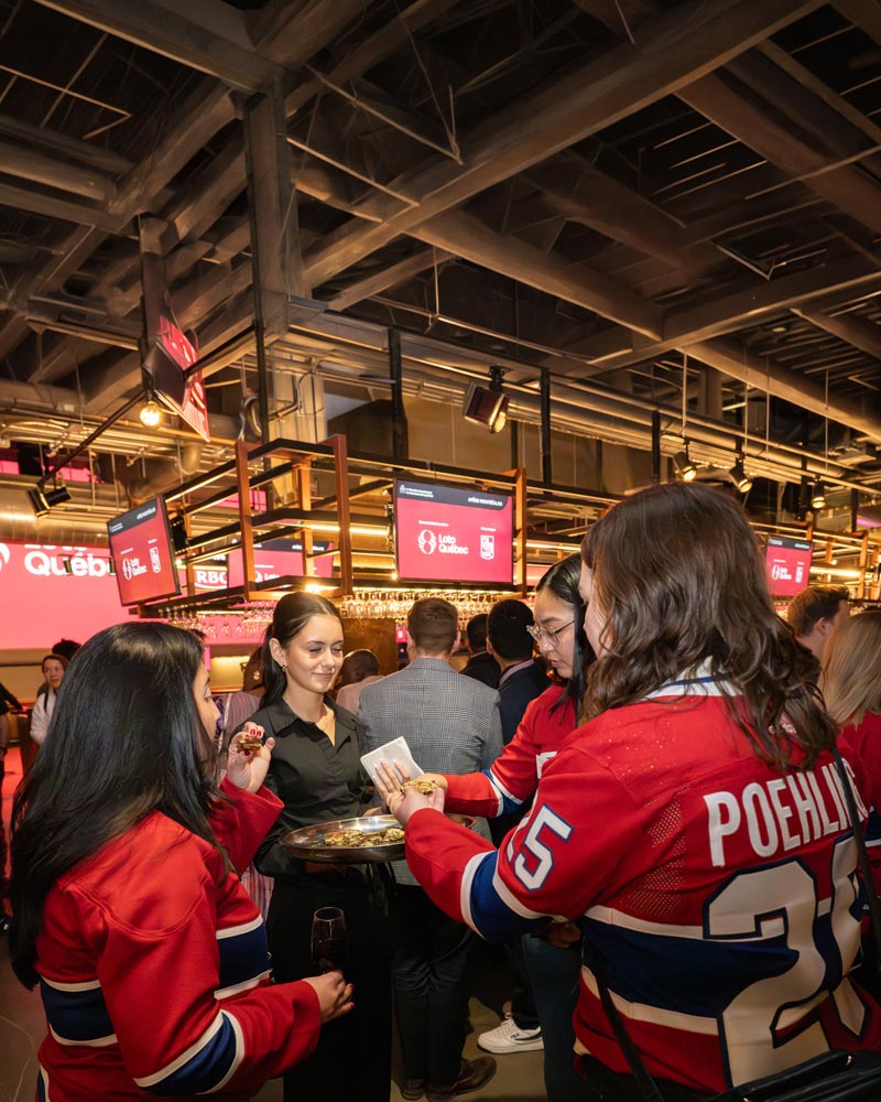 Participants à un événement de réseautage dans un espace de réception; plusieurs personnes portant des chandails de hockey échangent avec des invités, tandis qu’une personne sert des bouchées. Des écrans affichant des logos de partenaires sont visibles en arrière-plan.
