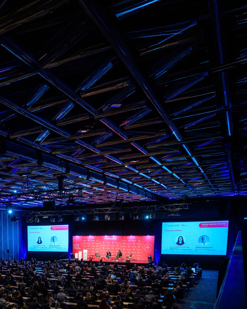 Vue d’ensemble d’une conférence de la Chambre de commerce du Montréal métropolitain; plusieurs participants sont réunis dans une grande salle face à une scène où se déroule une discussion, avec des écrans de présentation et un éclairage bleu et rouge.