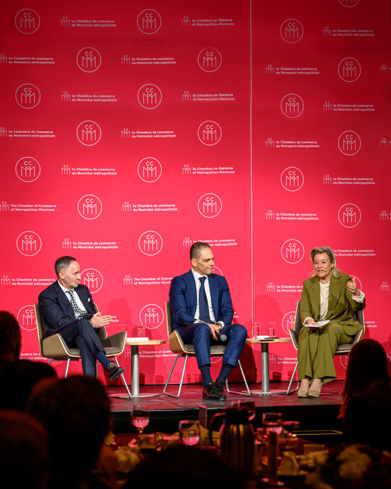 Discussion sur scène lors d’un événement de la Chambre de commerce du Montréal métropolitain; plusieurs panélistes participent à un échange devant un auditoire, sur fond rouge aux couleurs de la CCMM.
