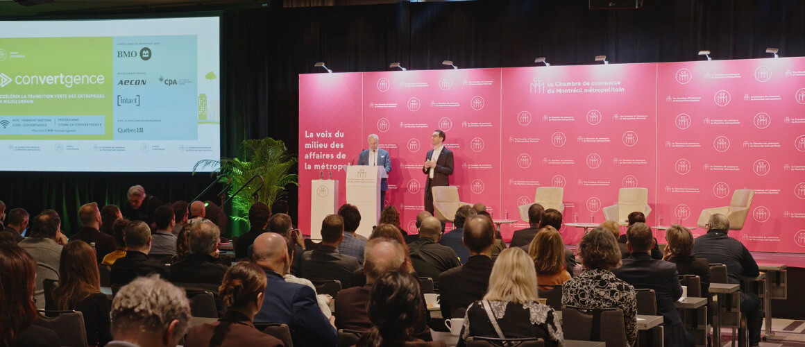 Two men in suits, one at a lectern, are speaking to an audience at a Chamber of Commerce of Metropolitan Montreal event. A large screen behind them displays the convergtence logo and a list of sponsors, while a pink wall features the Chamber of Commerce logo.