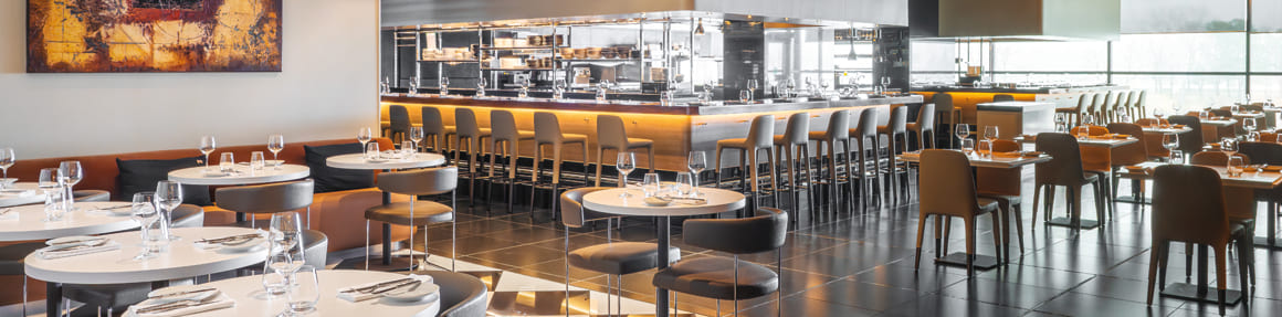 Modern restaurant dining room with set tables, aligned chairs and a long illuminated bar counter, in a bright space with contemporary design.
