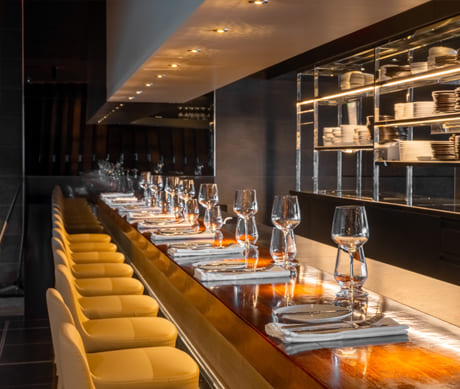 Long restaurant counter with aligned stools, tableware and glassware arranged on a wooden surface, in a dark and elegant space with open shelving in the background.