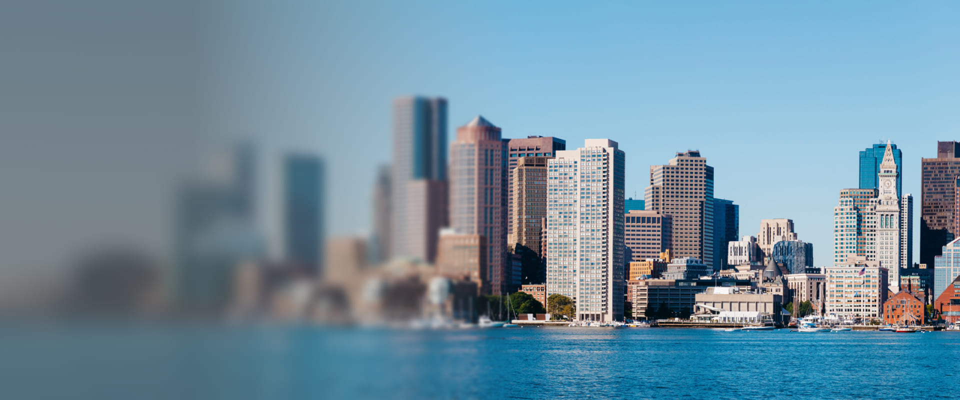 Panoramic view of downtown Boston featuring modern and historic skyscrapers along the waterfront under a clear blue sky.