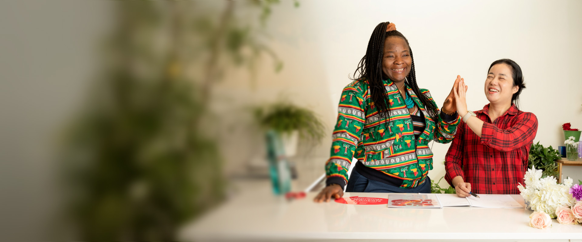 Deux personnes participent à une activité créative à une table; on voit des cartes, des fleurs et du matériel de bricolage, dans un espace lumineux avec des plantes en arrière-plan.