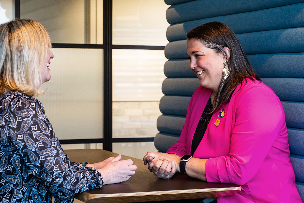 Deux femmes, souriantes et assises l'une en face de l'autre à une table sombre, semblent discuter. La femme à gauche, blonde, porte un haut à motifs sombres. La femme à droite, aux cheveux foncés, porte un blazer rose vif et un badge. L'arrière-plan flou montre une cloison rembourrée bleue.